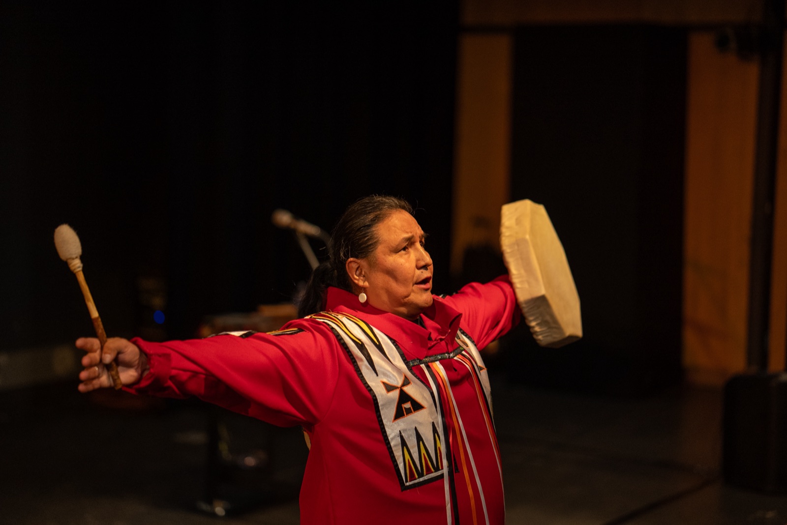 Walter White Bear on stage in red regalia, arms outstretched, holding a hand drum